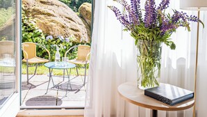 Une pièce dotée d’une porte en verre donnant sur un patio extérieur, une table ronde en bois sur laquelle repose un livre, un vase rempli de fleurs et des chaises en osier.