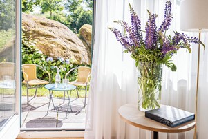 A room with a glass door leading to an outdoor patio, a round wooden table with a book, a vase with flowers, and wicker chairs.