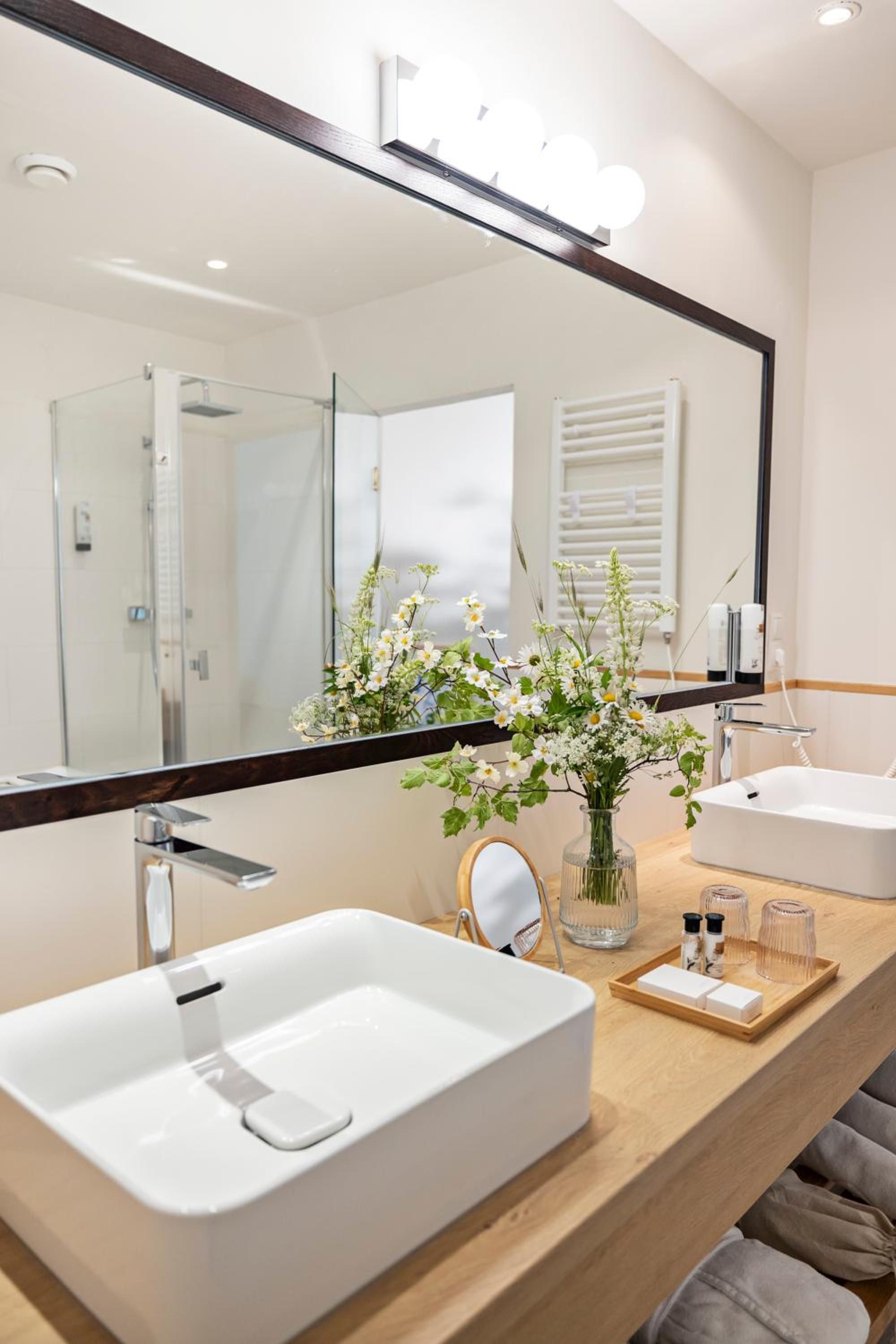 Un baño moderno con un espejo grande, dos lavamanos, una ducha y un florero con flores en el tocador.