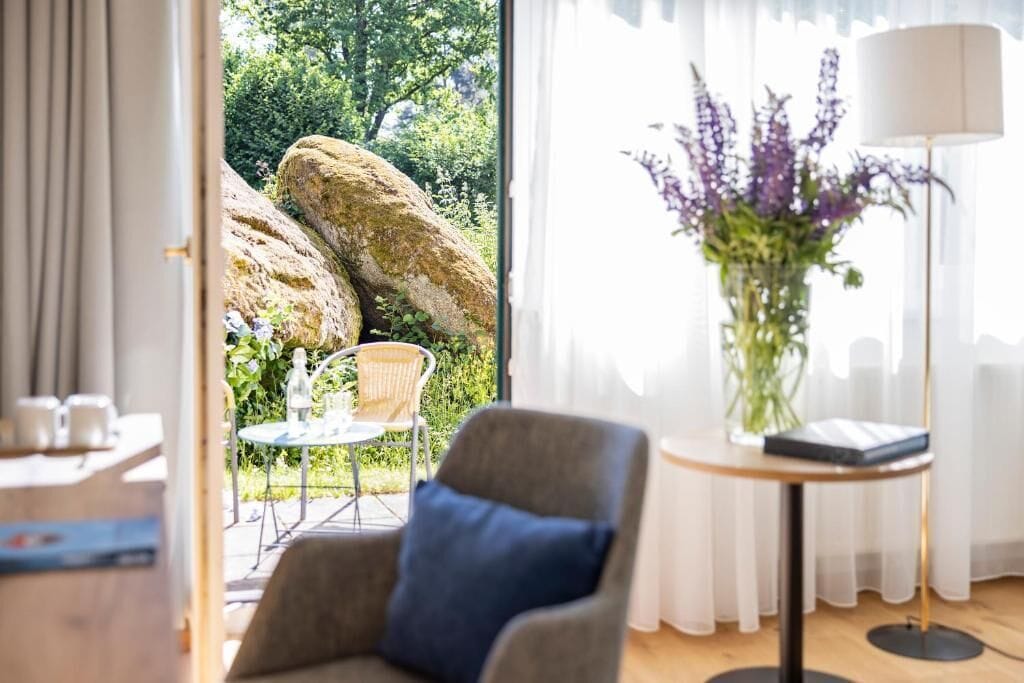 A room with a view of a garden, a chair, a table, and a lamp.