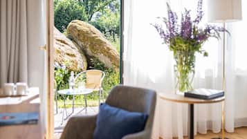 A room with a view of a garden, a chair, a table, and a lamp.
