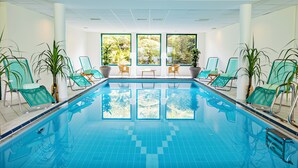 Une piscine intérieure à l’eau d’un bleu limpide, entourée de chaises longues et de plantes en pot, avec de grandes fenêtres donnant sur un paysage verdoyant.