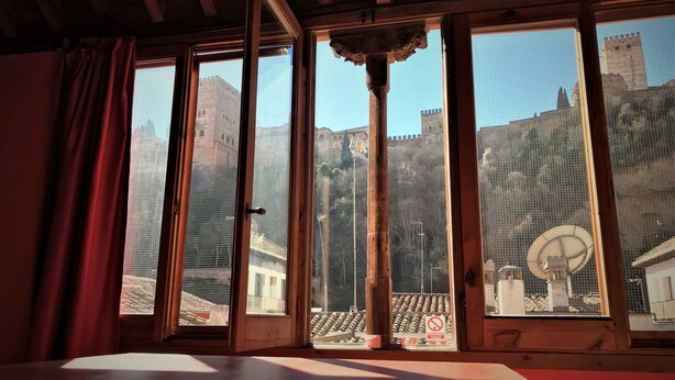 A room with large windows, wooden beams, and a view of a historic building. - White Nest (Granada)