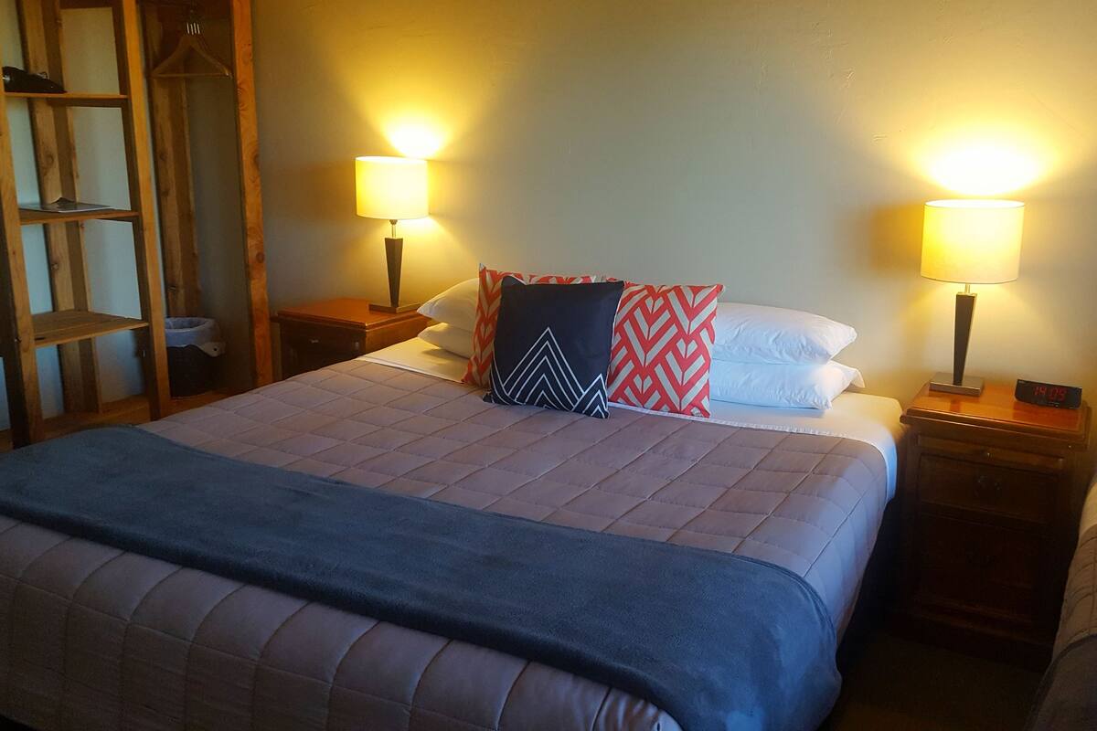 Uma cama de casal com um cobertor azul, duas mesas de cabeceira com luminárias e uma escada de madeira.