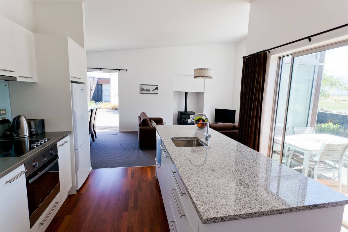 Uma cozinha moderna com bancada de granito, forno embutido e pia. Há uma área de jantar com mesa e cadeiras, e uma janela grande com vista para o exterior.