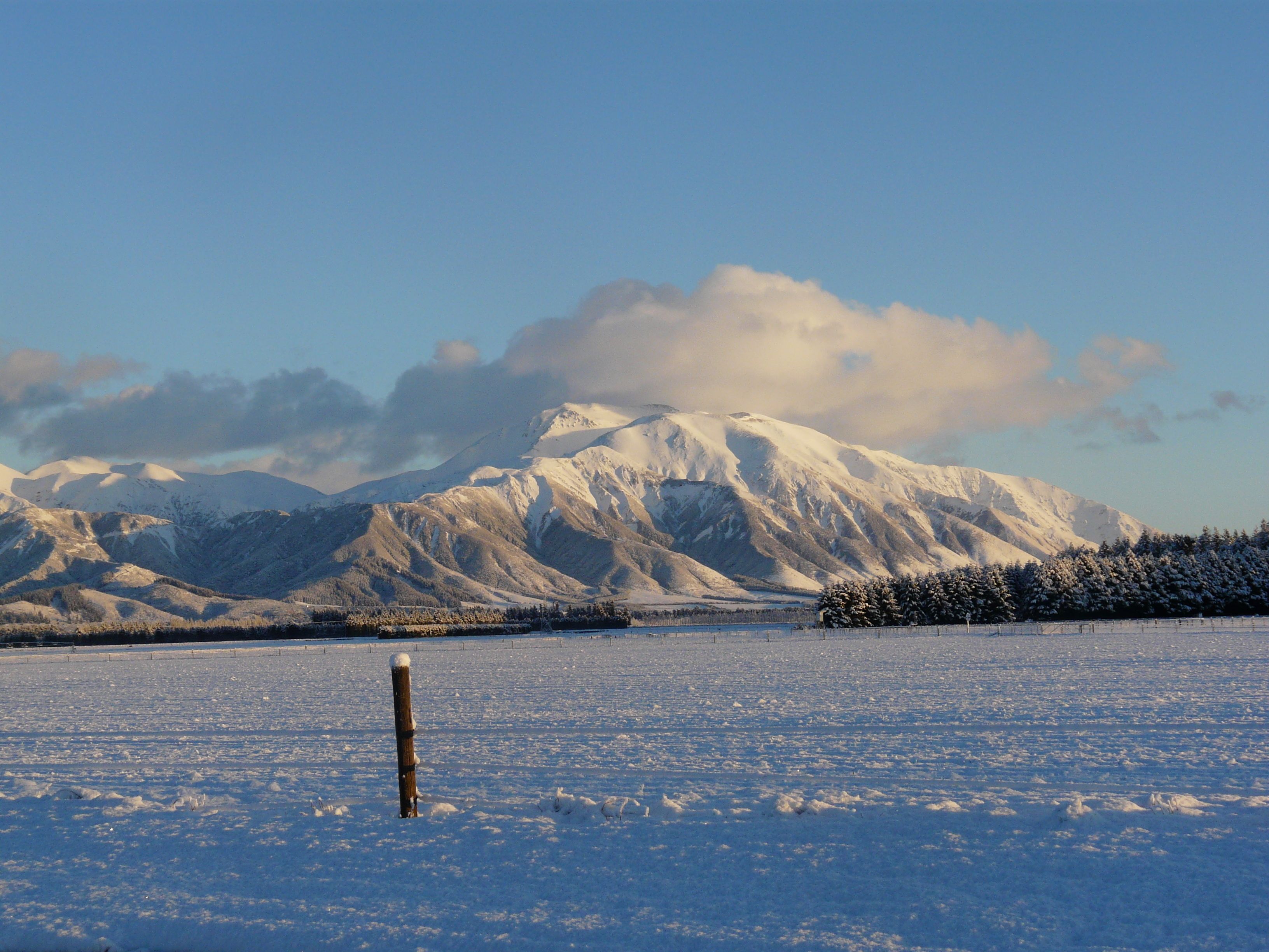 Schneelandschaft mit einem Gebirgszug im Hintergrund und einem hölzernen Pfosten im Vordergrund.