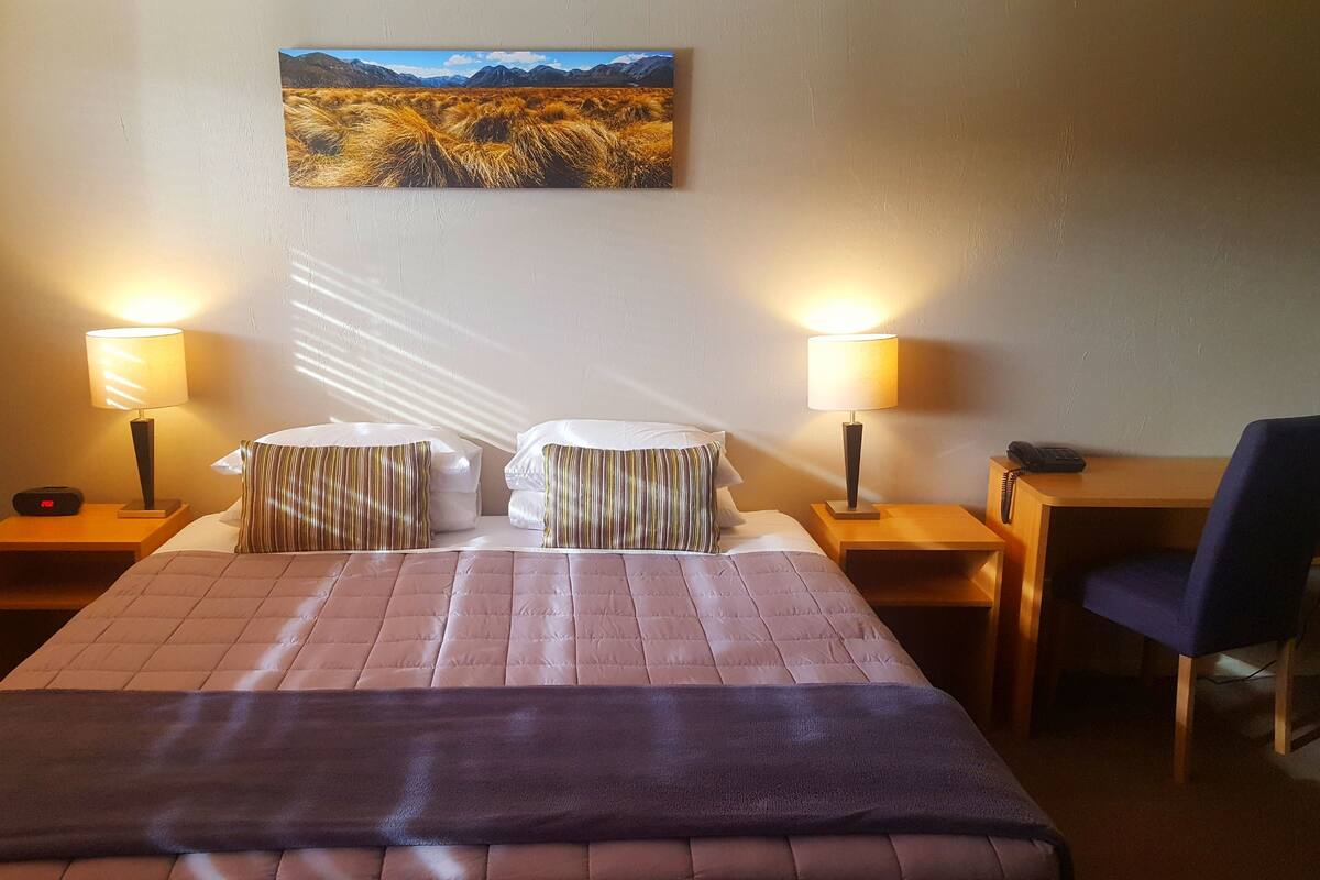 Quarto de hotel com cama, duas luminárias de cabeceira, uma escrivaninha com cadeira e um quadro na parede.