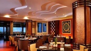 Un restaurante con una decoración moderna, que presenta una combinación de muebles de madera y metal, un amplio ventanal con vistas, y una pared decorativa con un emblema circular.