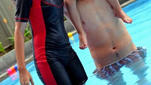 Una persona con traje de baño negro y rojo sostiene a un niño en un flotador azul sobre una piscina.