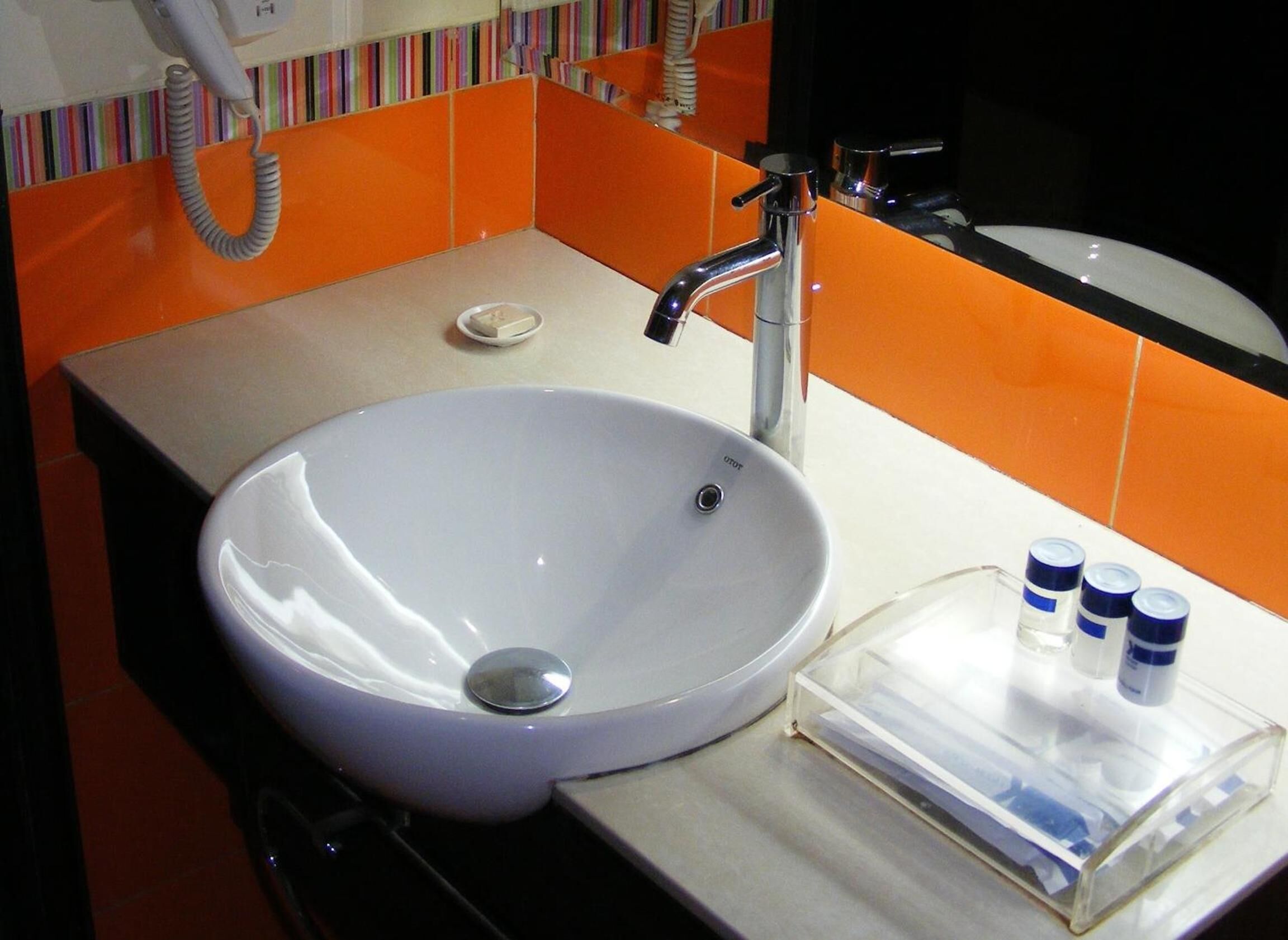 Un bagno con un lavandino bianco, un rubinetto cromato e uno specchio. Le pareti sono rivestite di piastrelle arancioni ed è presente un portascopevoli a righe.
