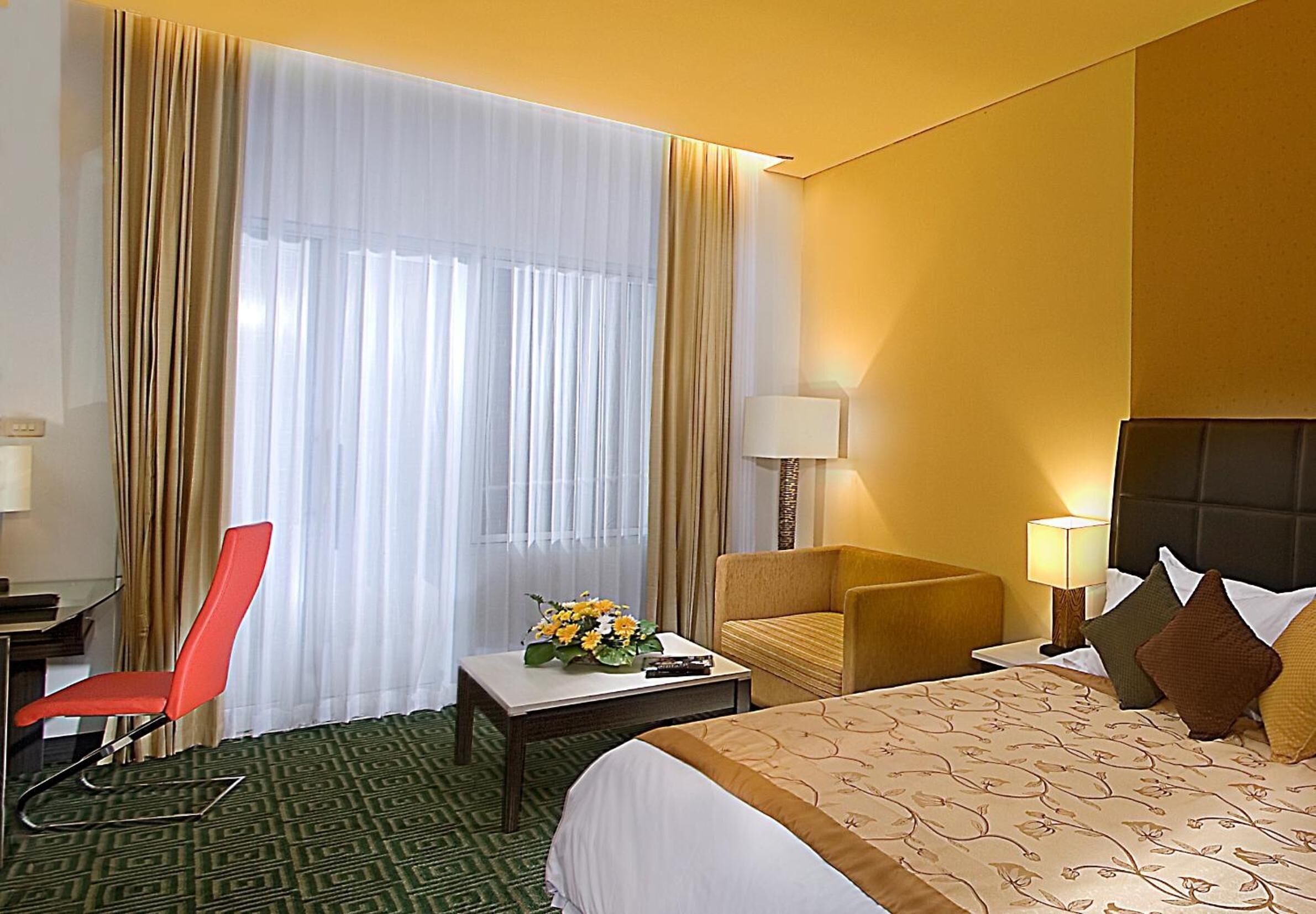Habitación de hotel con cama, escritorio, silla, lámpara y ventana con cortinas.