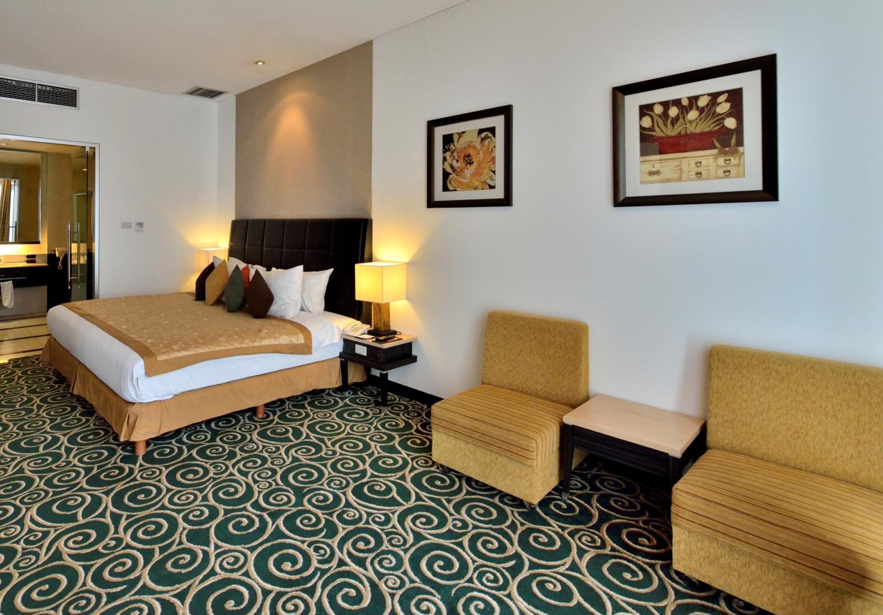 A hotel room with a bed, two bedside tables, a chair, a sofa, a patterned carpet, and two framed artworks on the wall.