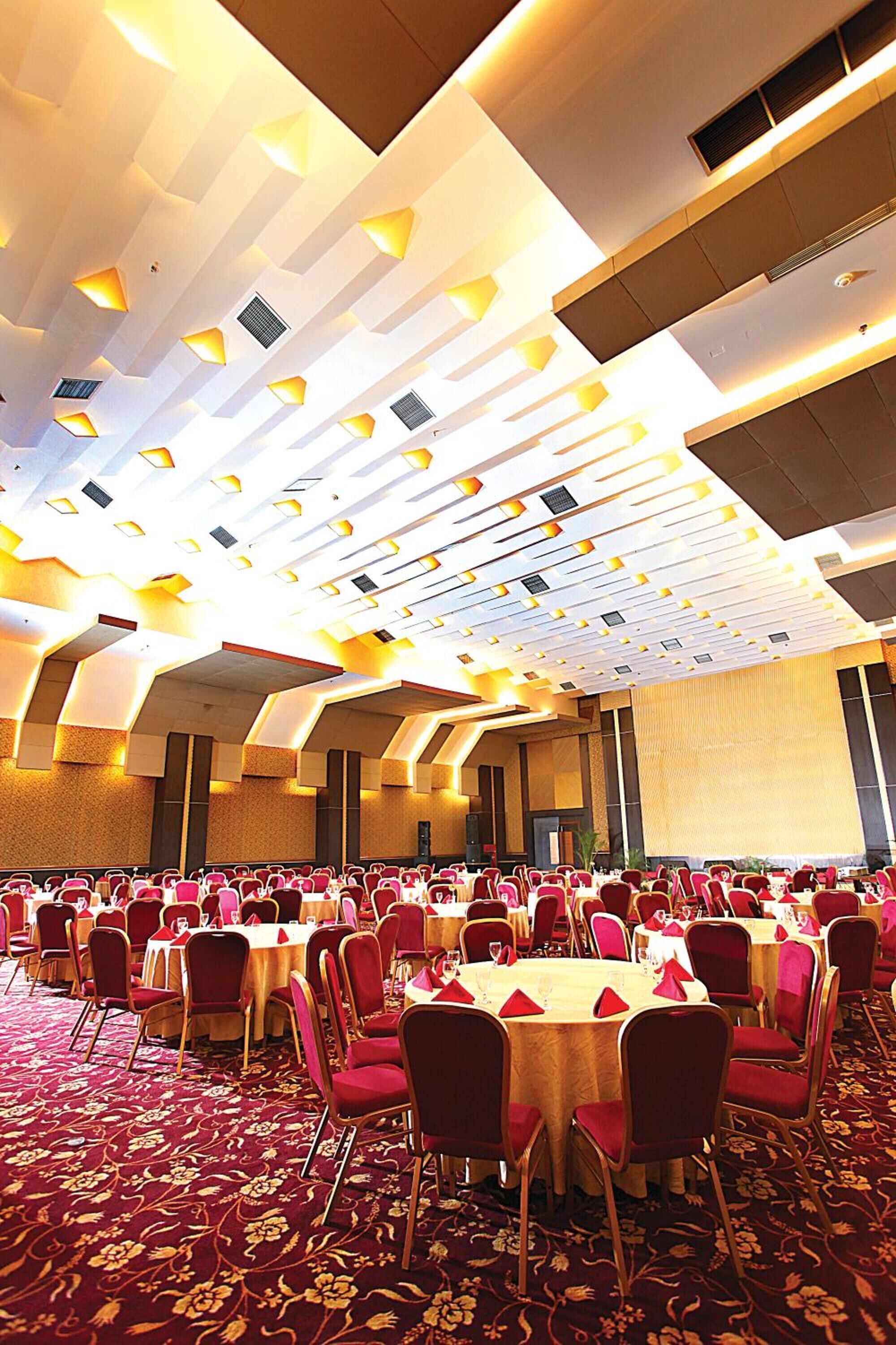 Une grande salle de banquet avec des tables rondes et des chaises rouges, agrémentée d’un tapis à motifs et d’un plafond au design moderne.