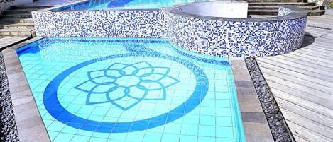 Piscina com padrão de mosaico azul, rodeada por um deck de pedra e um edifício branco com uma placa.