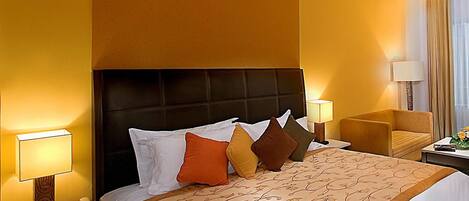 A hotel room with a large bed, a headboard, two bedside tables with lamps, a chair, and a nightstand with a phone and a notepad.