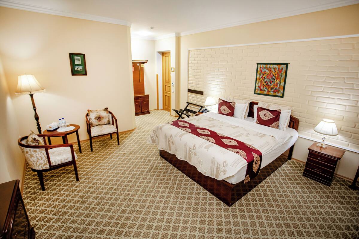A hotel room with a large bed, two chairs, a small table, a nightstand, and a wall-mounted lamp.