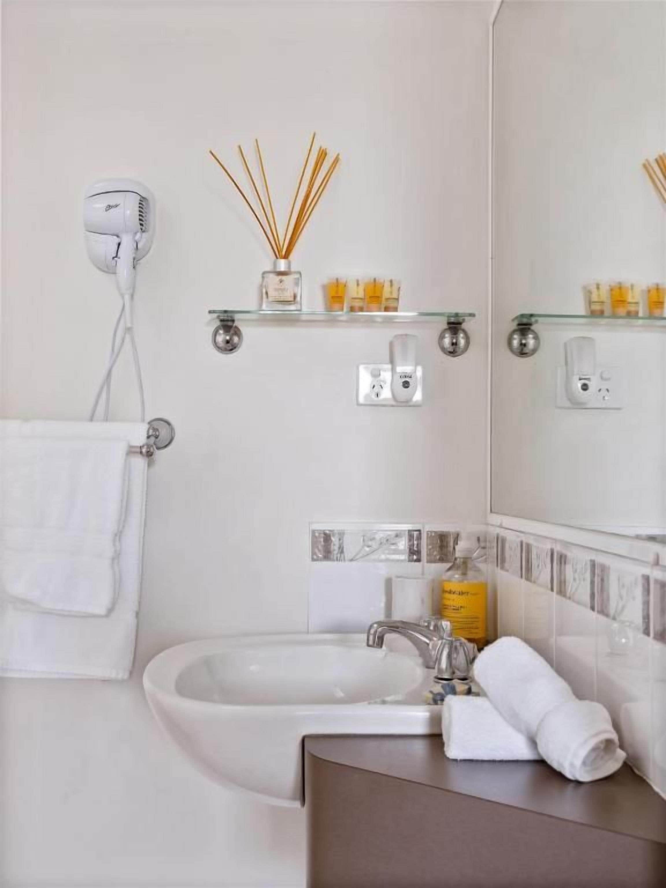 Baño con lavamanos, espejo y secador de pelo en la pared. Se observan estantes con artículos de aseo personal y un perchero.
