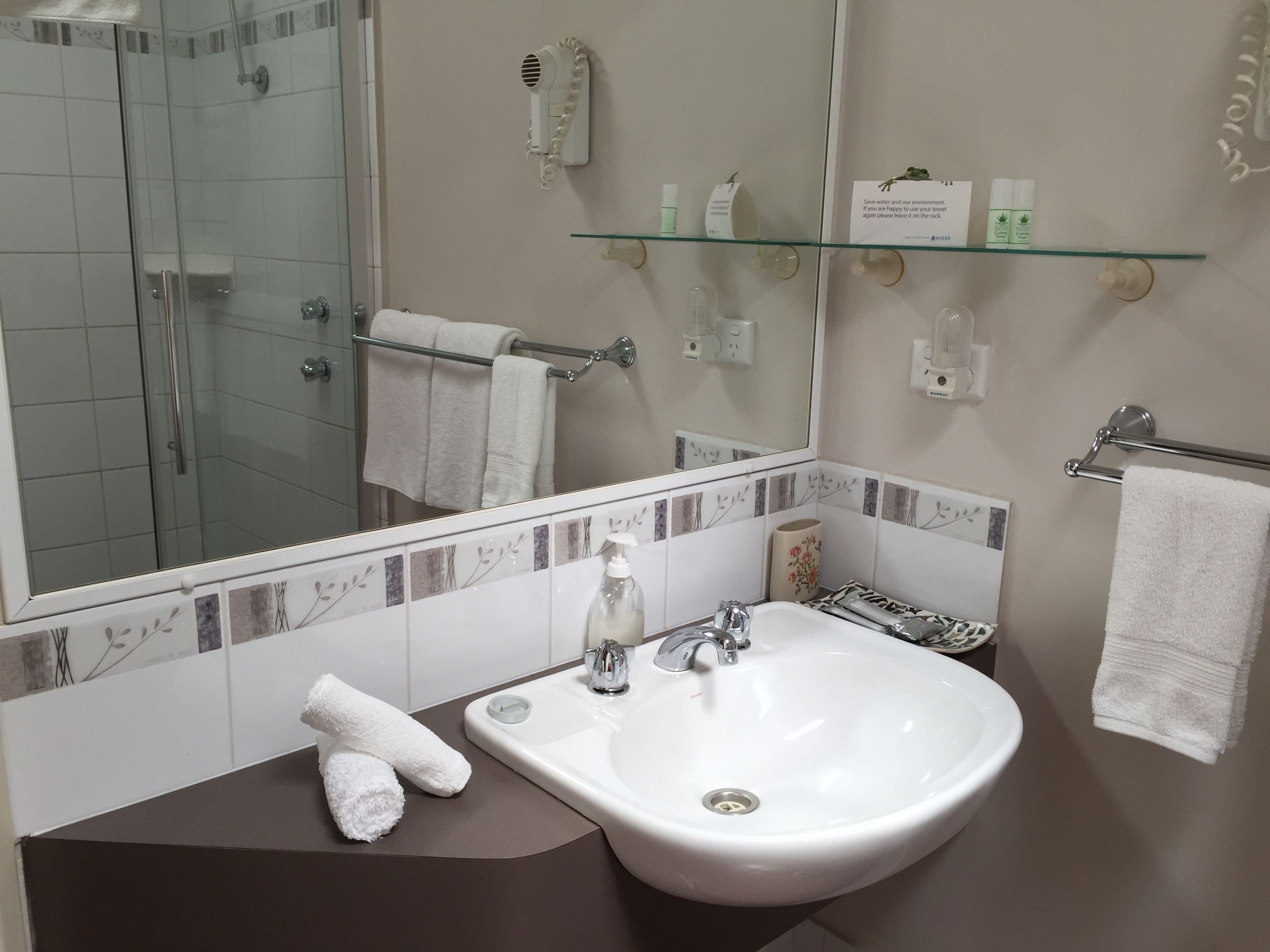 A modern bathroom with a glass shower enclosure, a white sink with a single faucet, and a glass shelf above the sink holding toiletries.