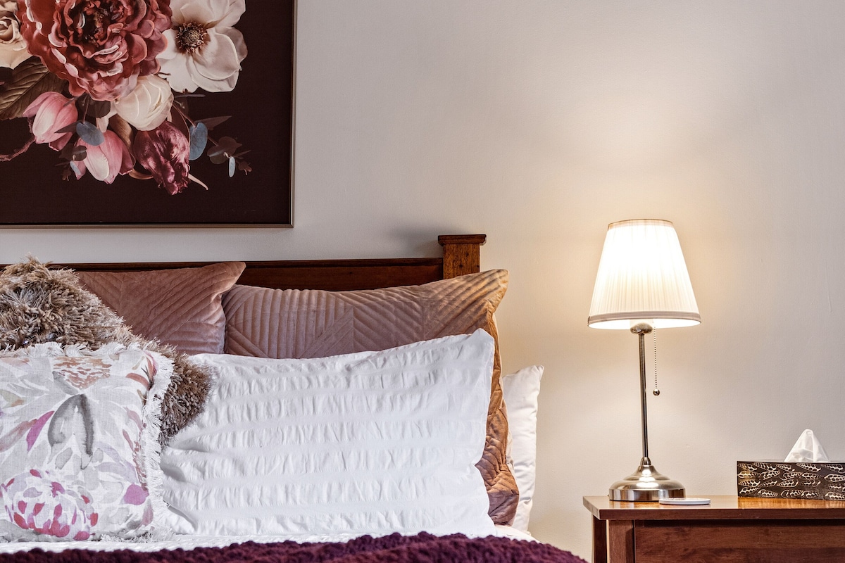 Una cama bien hecha con una colcha blanca y varias almohadas, una mesita de noche con una lámpara y una obra de arte floral en la pared.