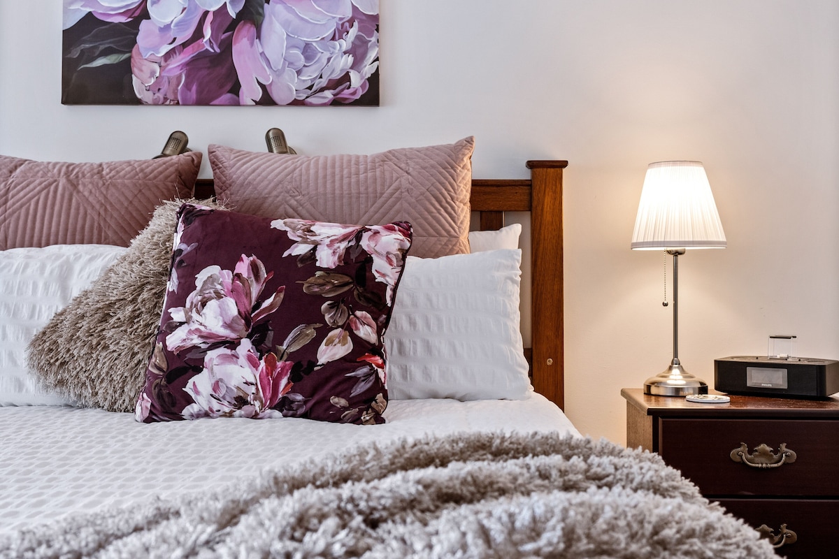 Una cama bien hecha con almohadas florales y con textura, una lámpara de noche y una mesita de noche de madera con una radio.