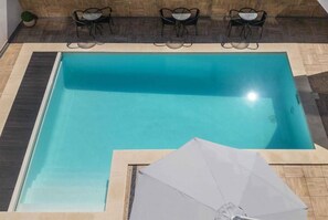 A rectangular indoor swimming pool with a clear blue water surface, surrounded by a tiled deck. There are three black metal chairs and a small round table on the deck, and a white umbrella is placed on the deck near the pool.