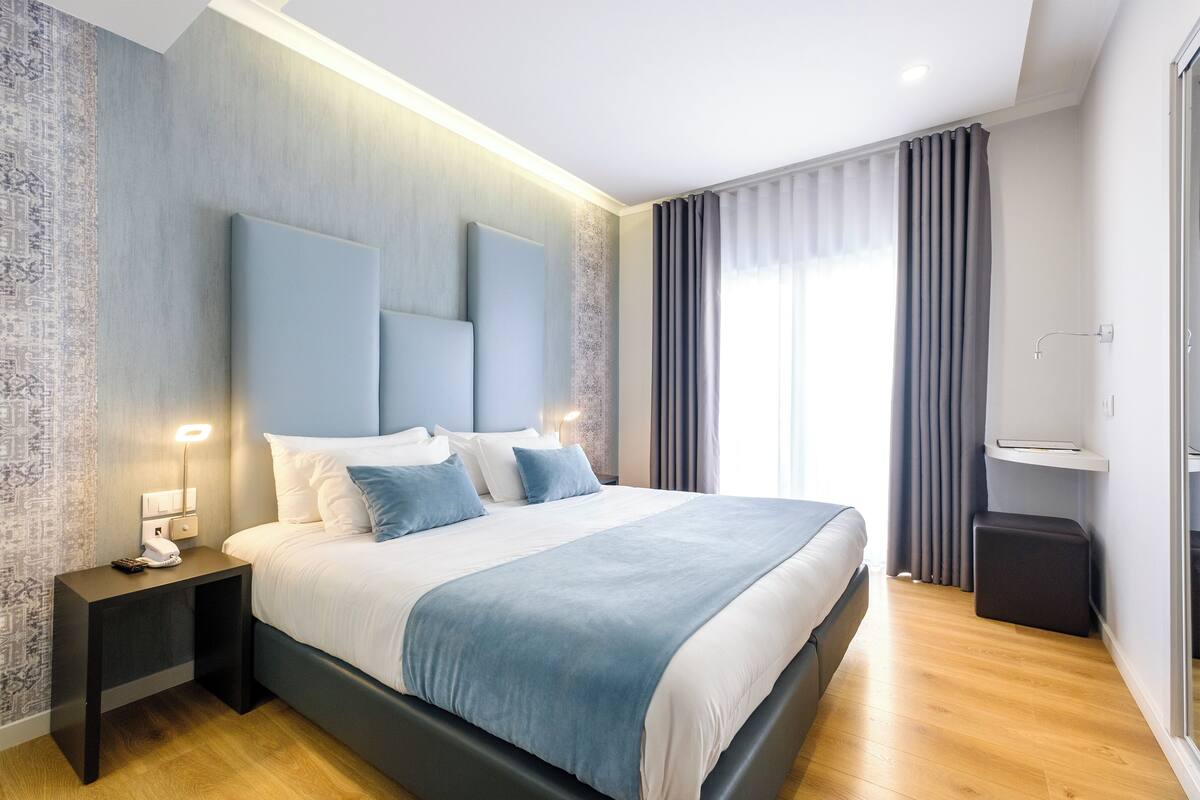 A modern hotel room with a large bed, bedside tables, a nightstand, a built-in TV shelf, and a window with curtains.