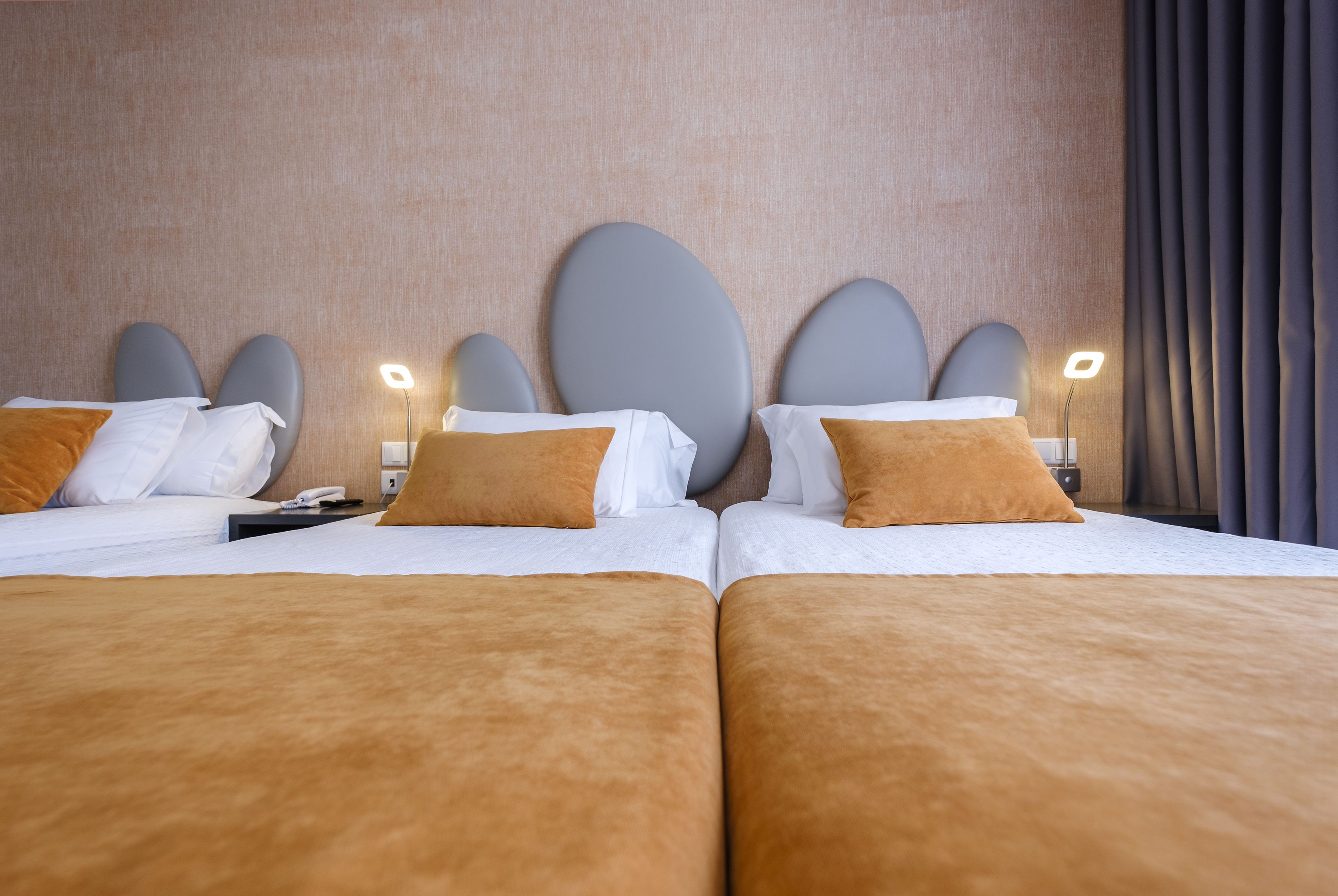 Quarto de hotel com duas camas, cada uma com cabeceira, roupa de cama branca e mantas marrons. Há mesinhas de cabeceira com luminárias e um telefone em uma das camas.