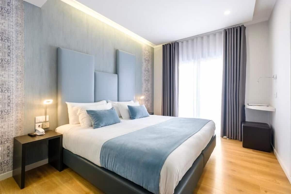A modern hotel room with a large bed, bedside tables, a nightstand, a small seating area, and a window with curtains.