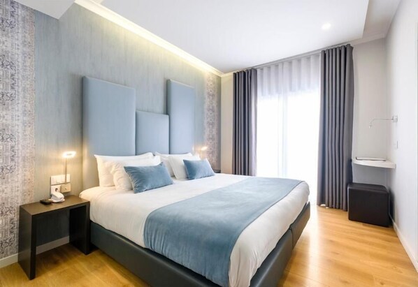 A modern hotel room with a large bed, bedside tables, a nightstand, a small seating area, and a window with curtains.