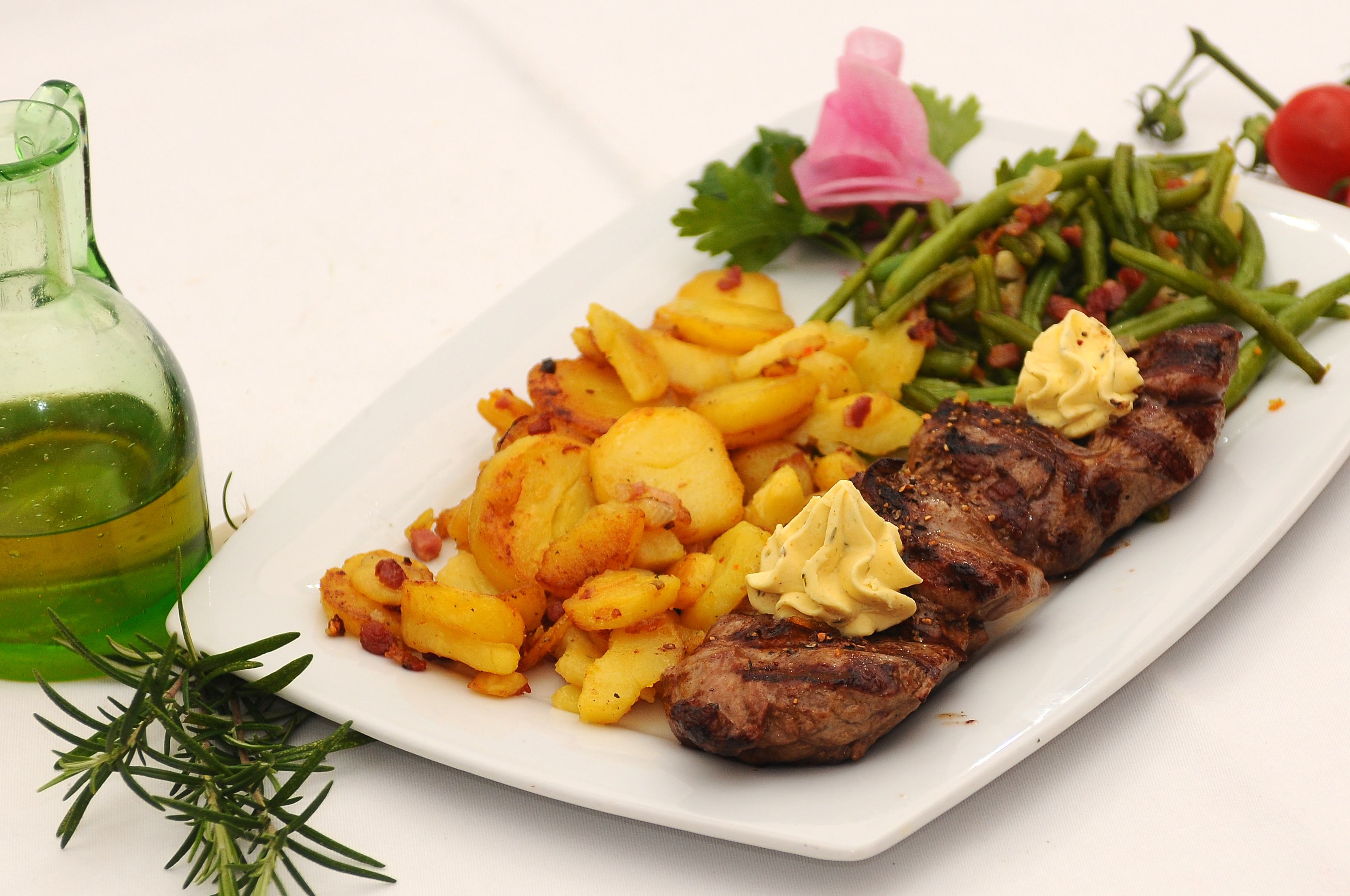 A plate with grilled meat, roasted potatoes, and green beans, accompanied by a bottle of oil and a sprig of rosemary.
