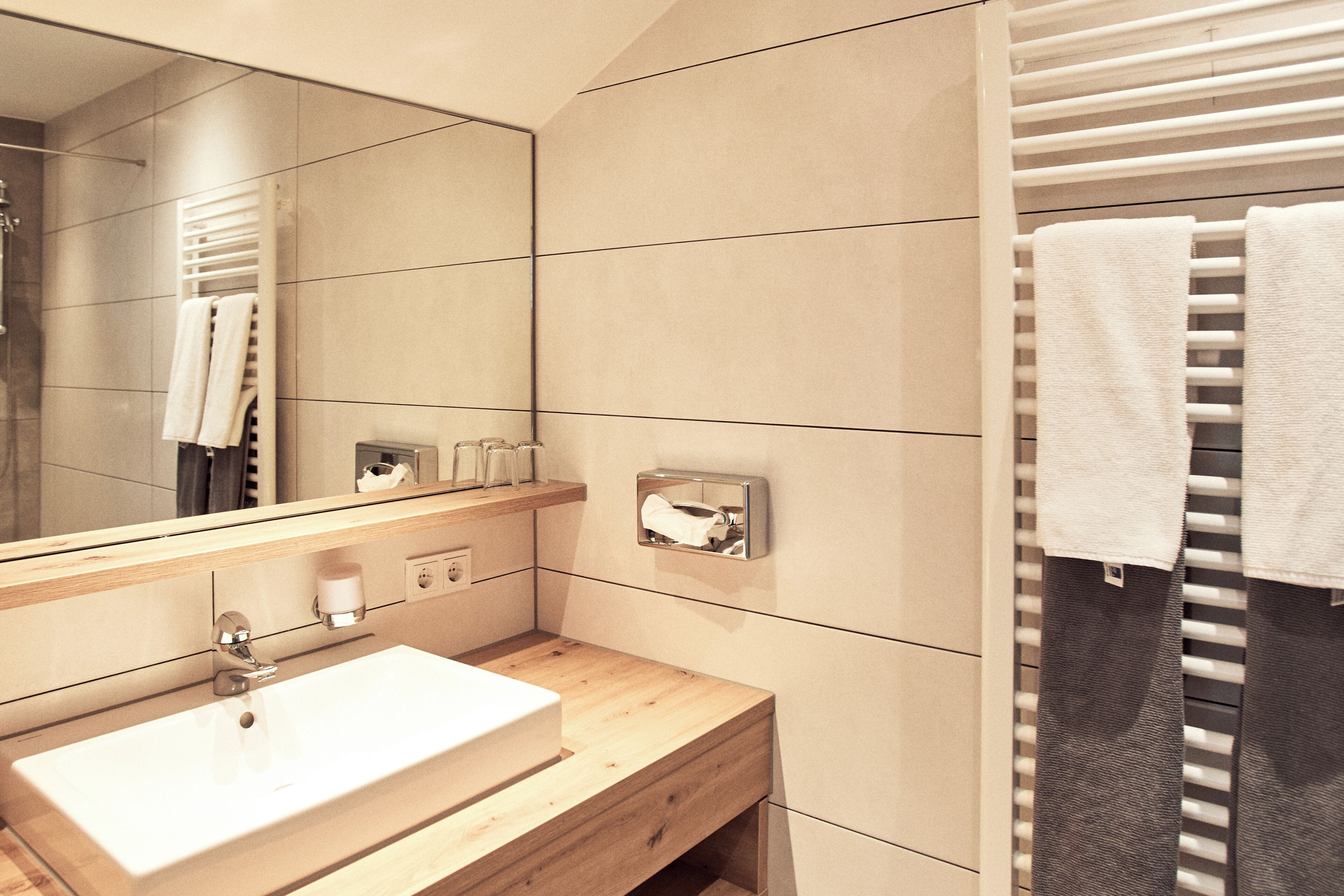 Un baño moderno con un espejo grande, un lavamanos blanco, tocador de madera, toallero y dispensador de toallas.