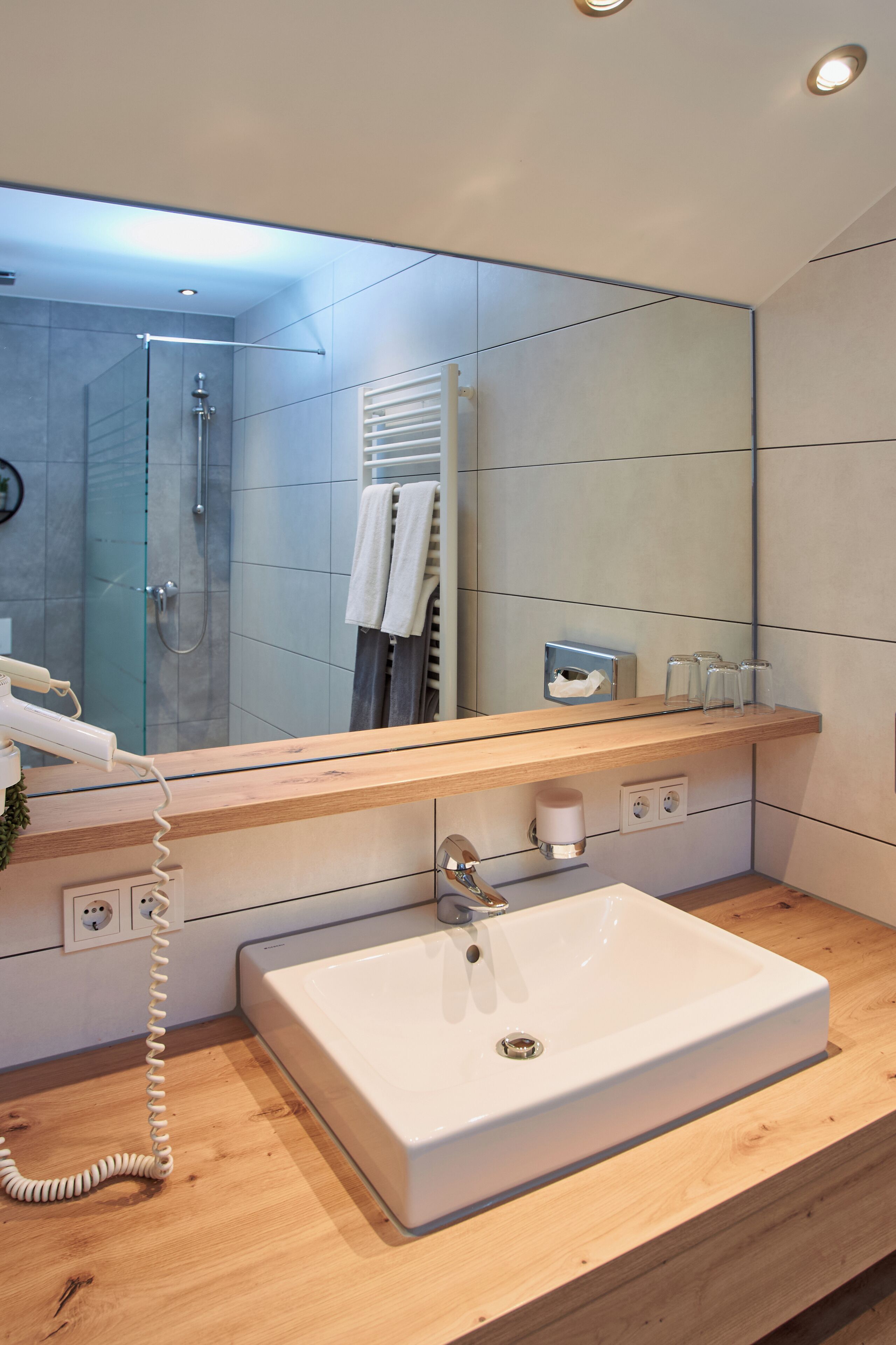 A modern bathroom with a large mirror, a wooden vanity, a white sink, and a glass-enclosed shower.