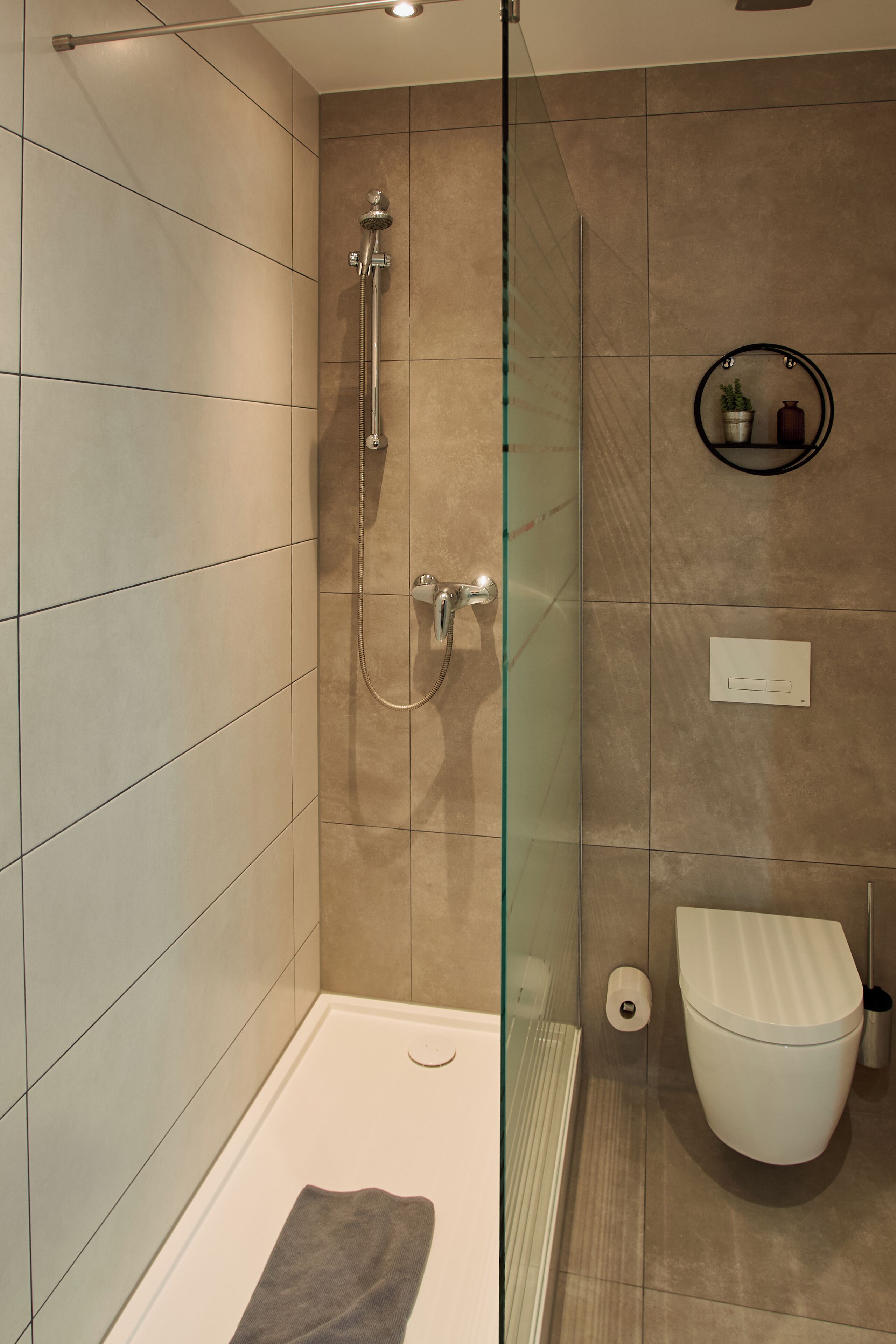 Une salle de bain moderne avec une douche à parois de verre, des toilettes blanches et une petite étagère contenant des articles de toilette.