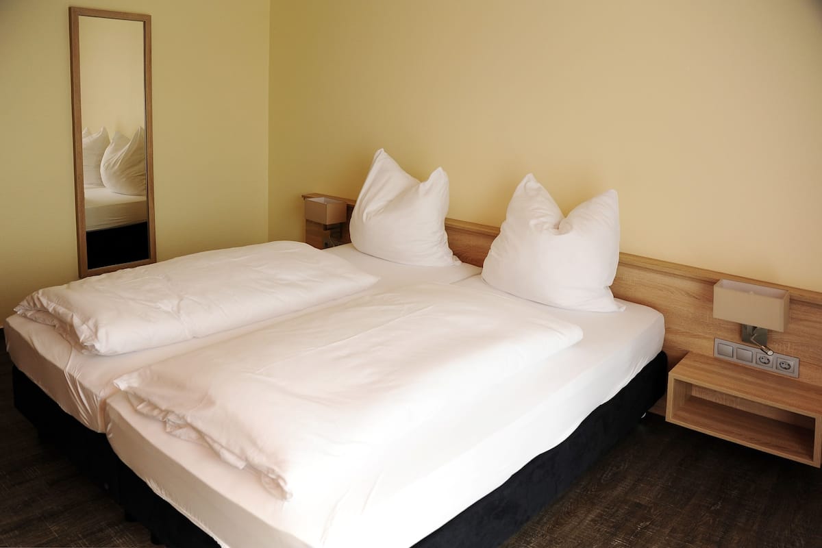 A neatly made bed with white linens and pillows, a wooden nightstand with a lamp, and a mirror on the wall.