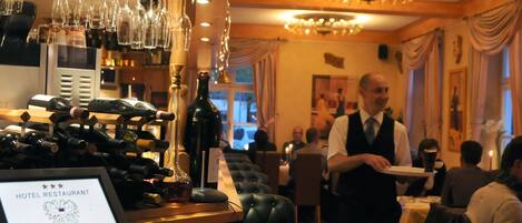 A hotel restaurant interior with a bar area, wine bottles, and a waiter serving guests.