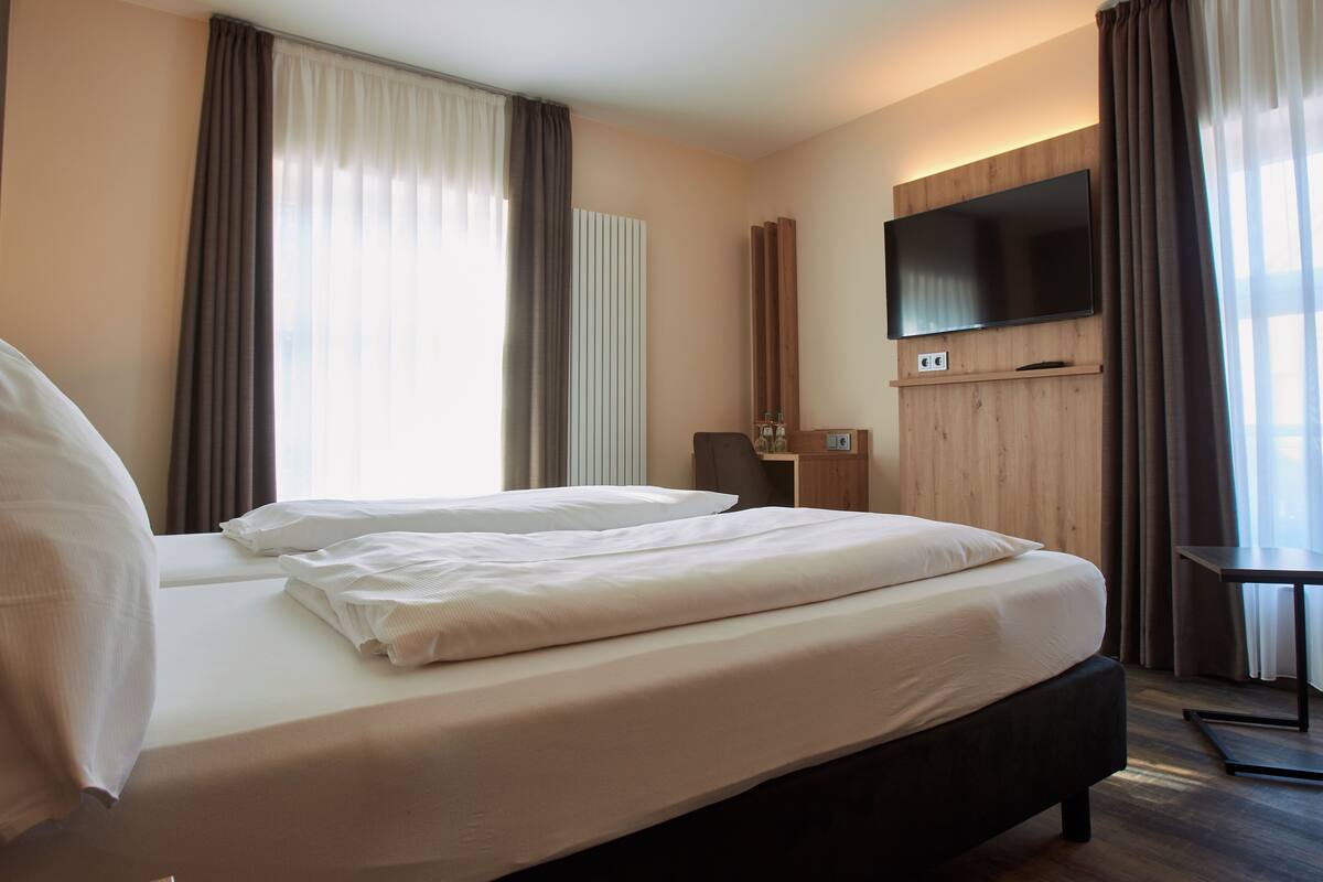 A hotel room with a large bed, a flat-screen TV mounted on the wall, and a desk with a chair.