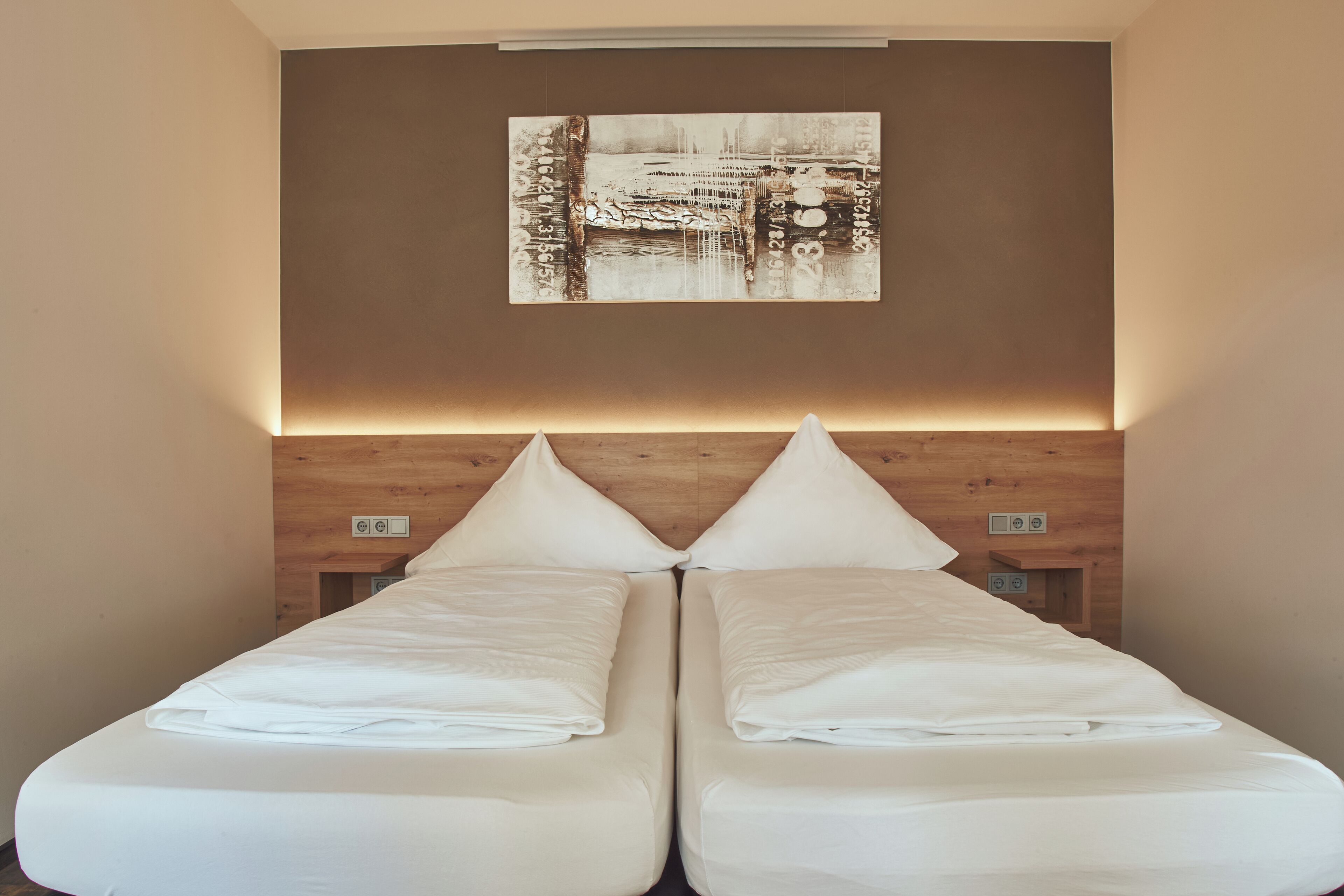A double bed with white linens, a headboard with a light wood finish, and a wall-mounted artwork above the bed.