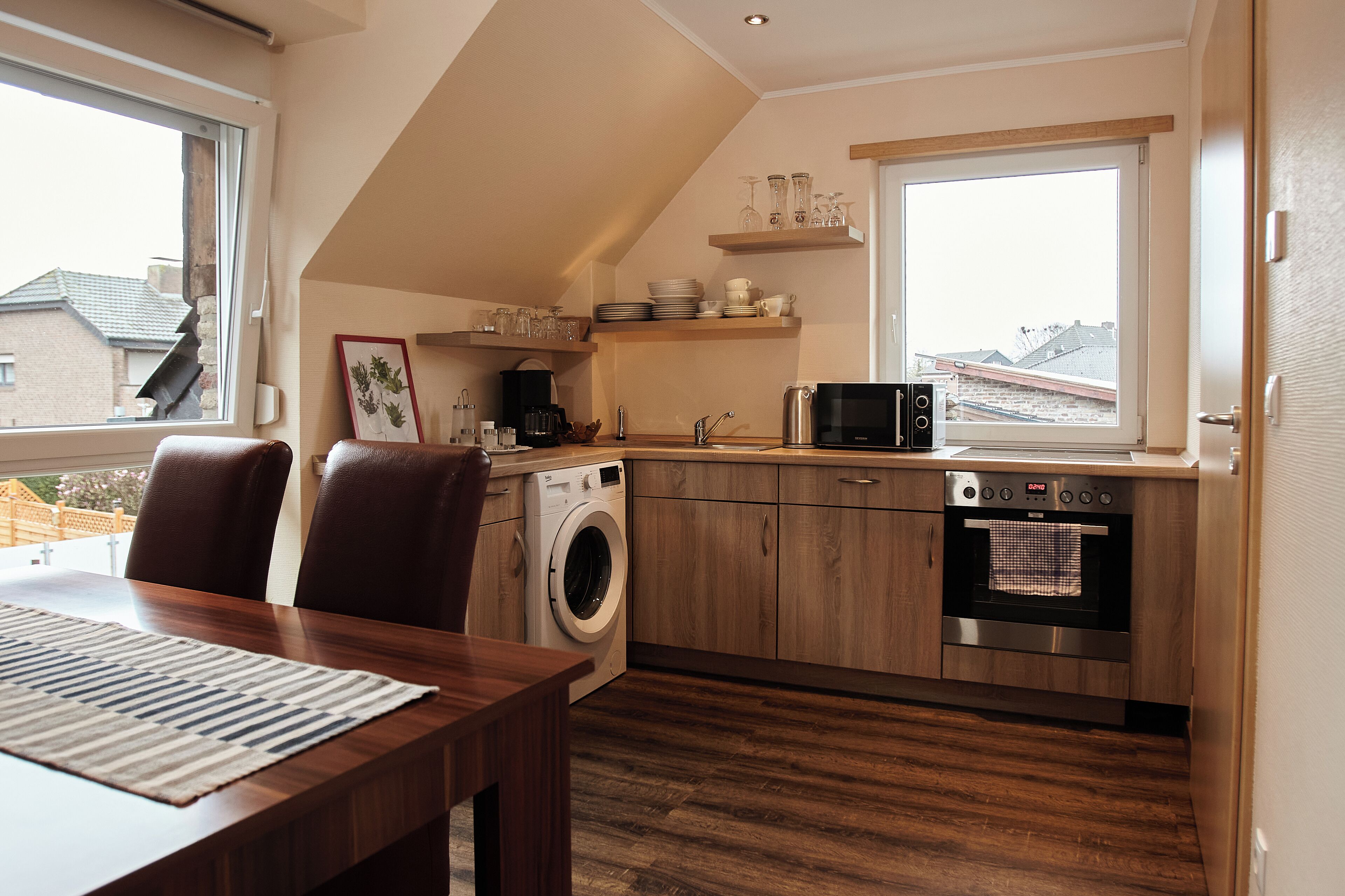 A compact kitchen with a washing machine, oven, and microwave. There is a dining table with chairs and a window providing a view of buildings outside.
