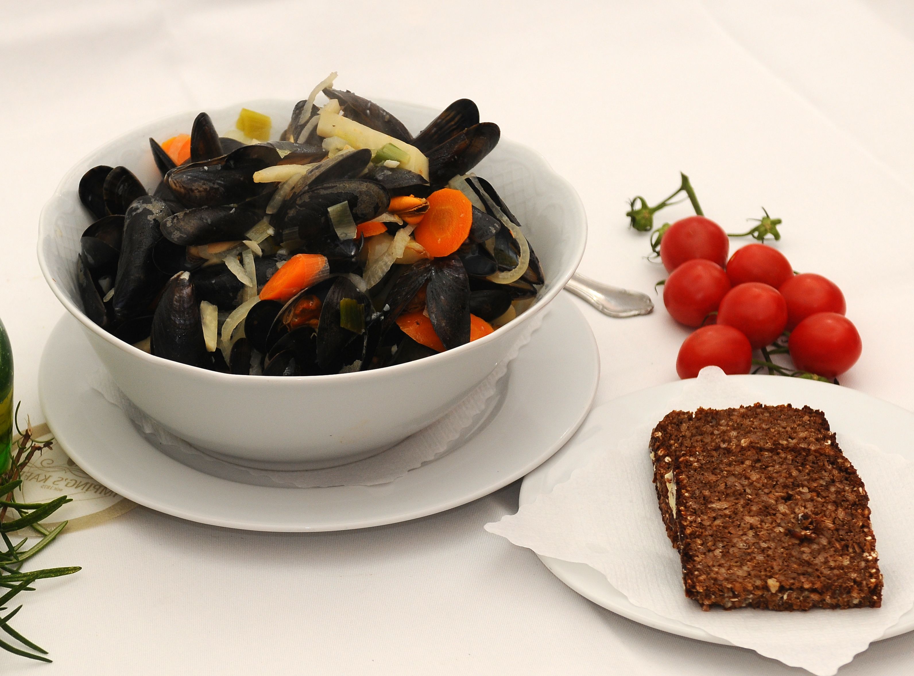 A bowl of mussels with vegetables on a white plate, accompanied by a slice of dark bread and cherry tomatoes on the side.