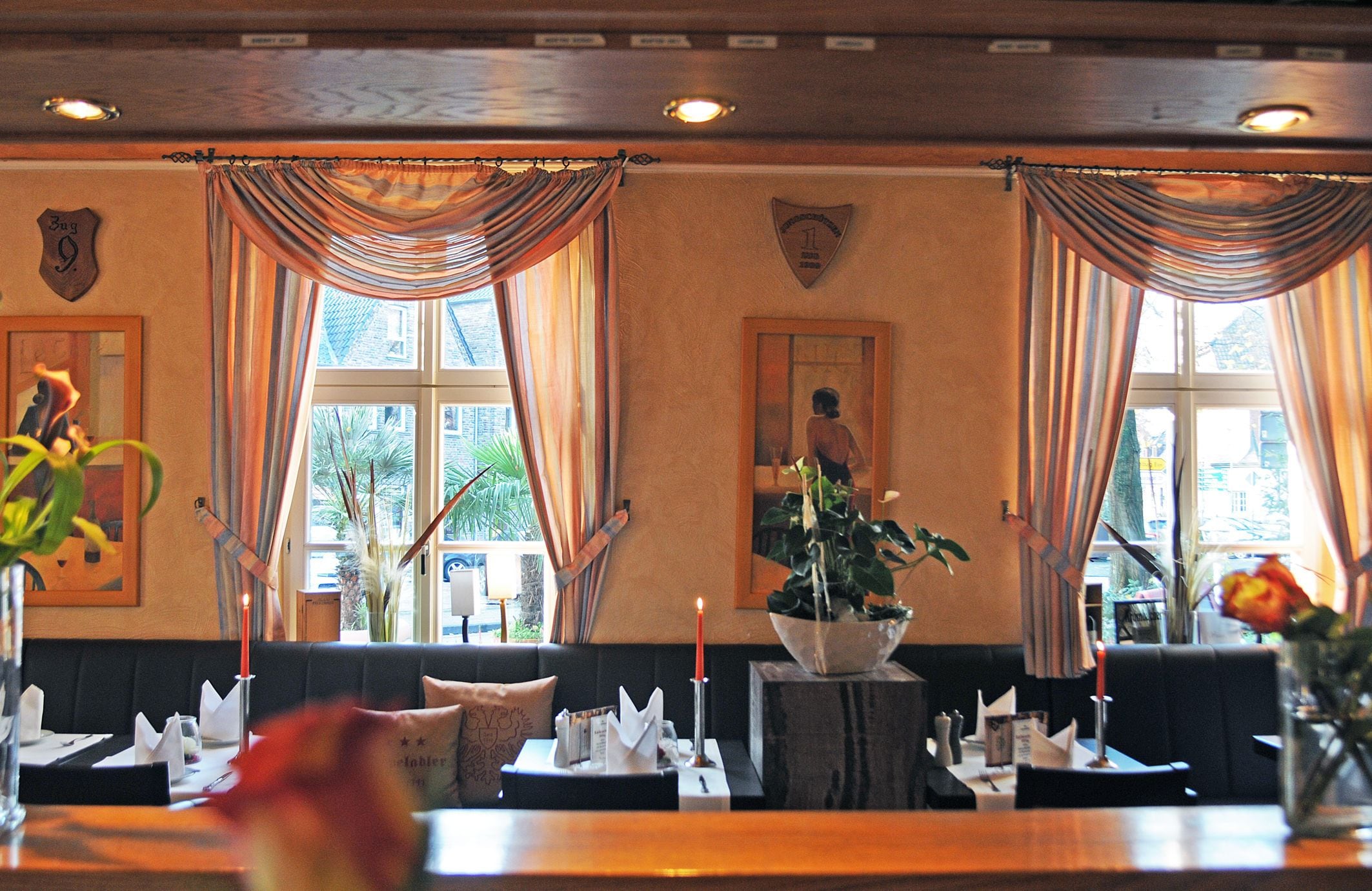 A restaurant with tables set for dining, a waiter in the background, and large windows with curtains.