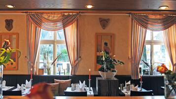 A restaurant with tables set for dining, a waiter in the background, and large windows with curtains.