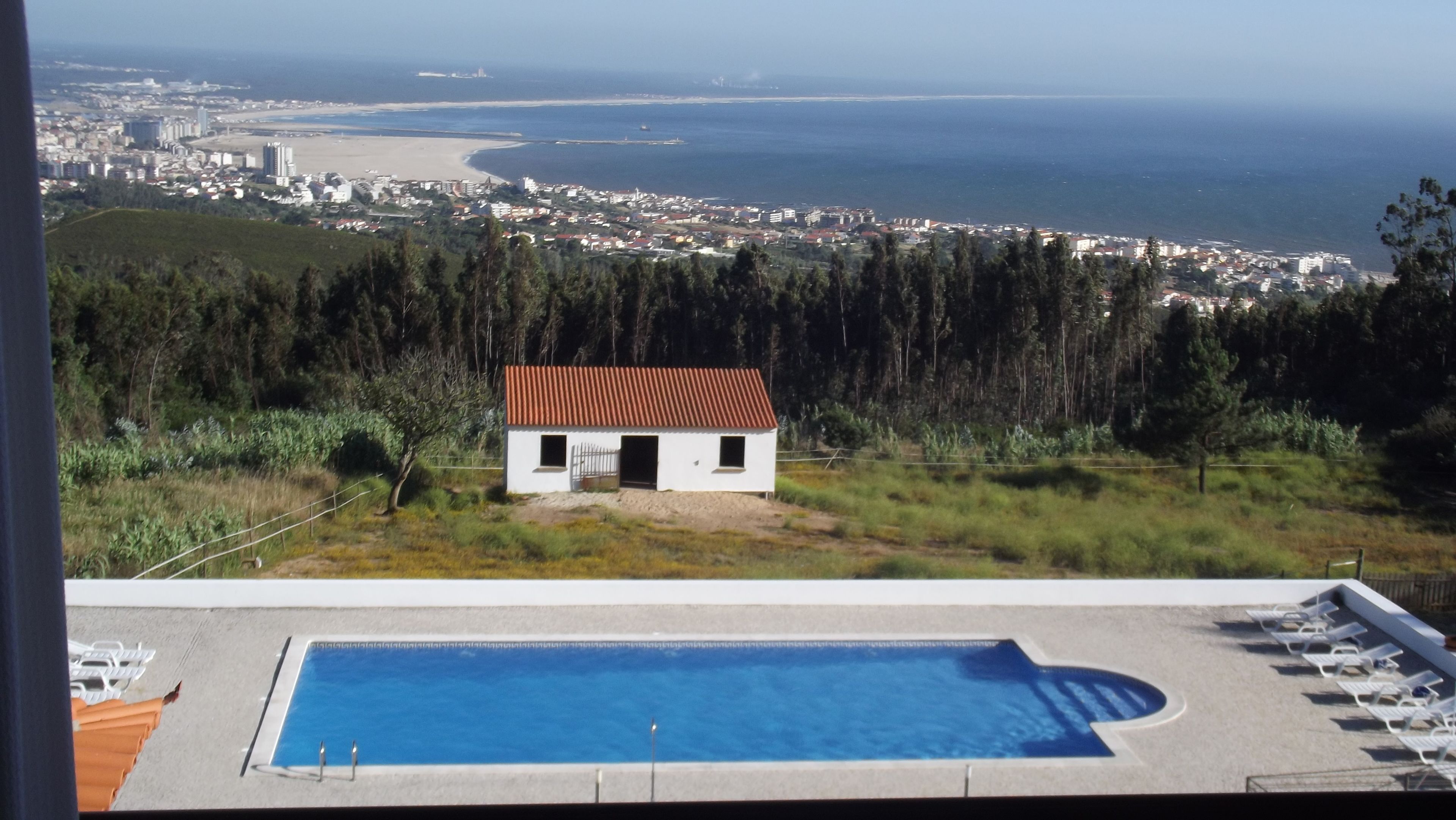 Vista da piscina com um pequeno edifício branco e teto vermelho, rodeado por vegetação, com vista para uma paisagem urbana costeira.