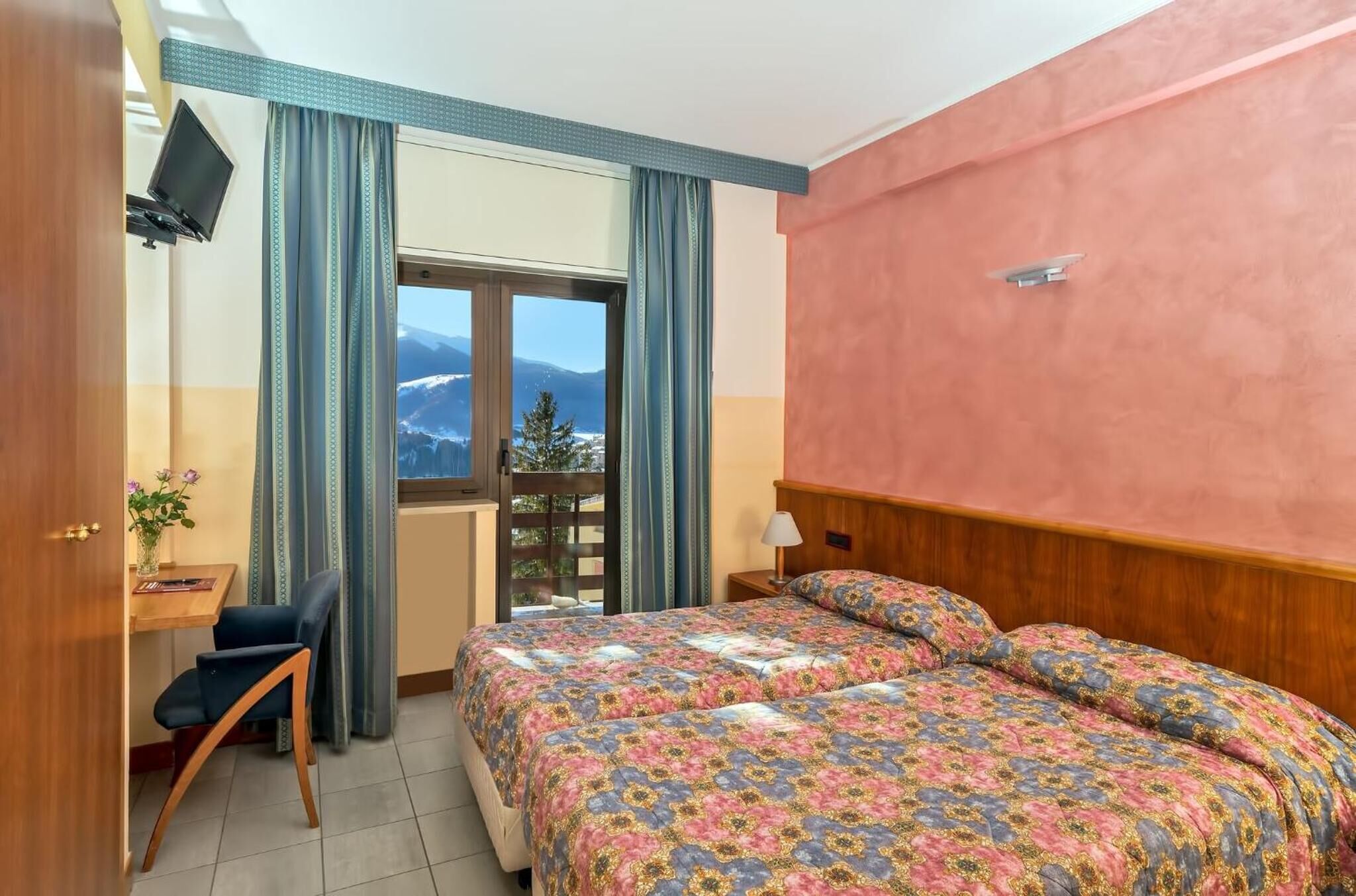 Una camera da letto con un letto, una scrivania e una sedia. Si apre una finestra con vista sulle montagne.