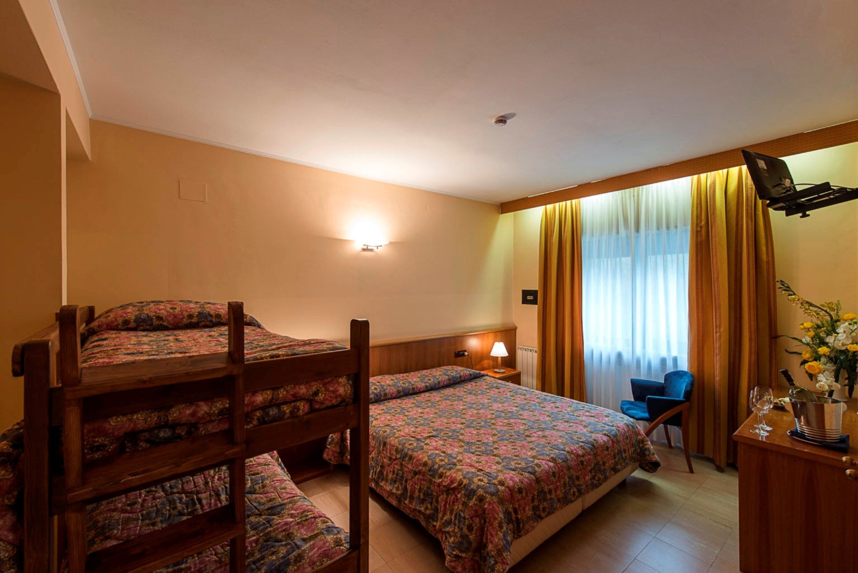 A hotel room with a bunk bed, a single bed, a desk with a chair, a lamp, and a window with curtains.