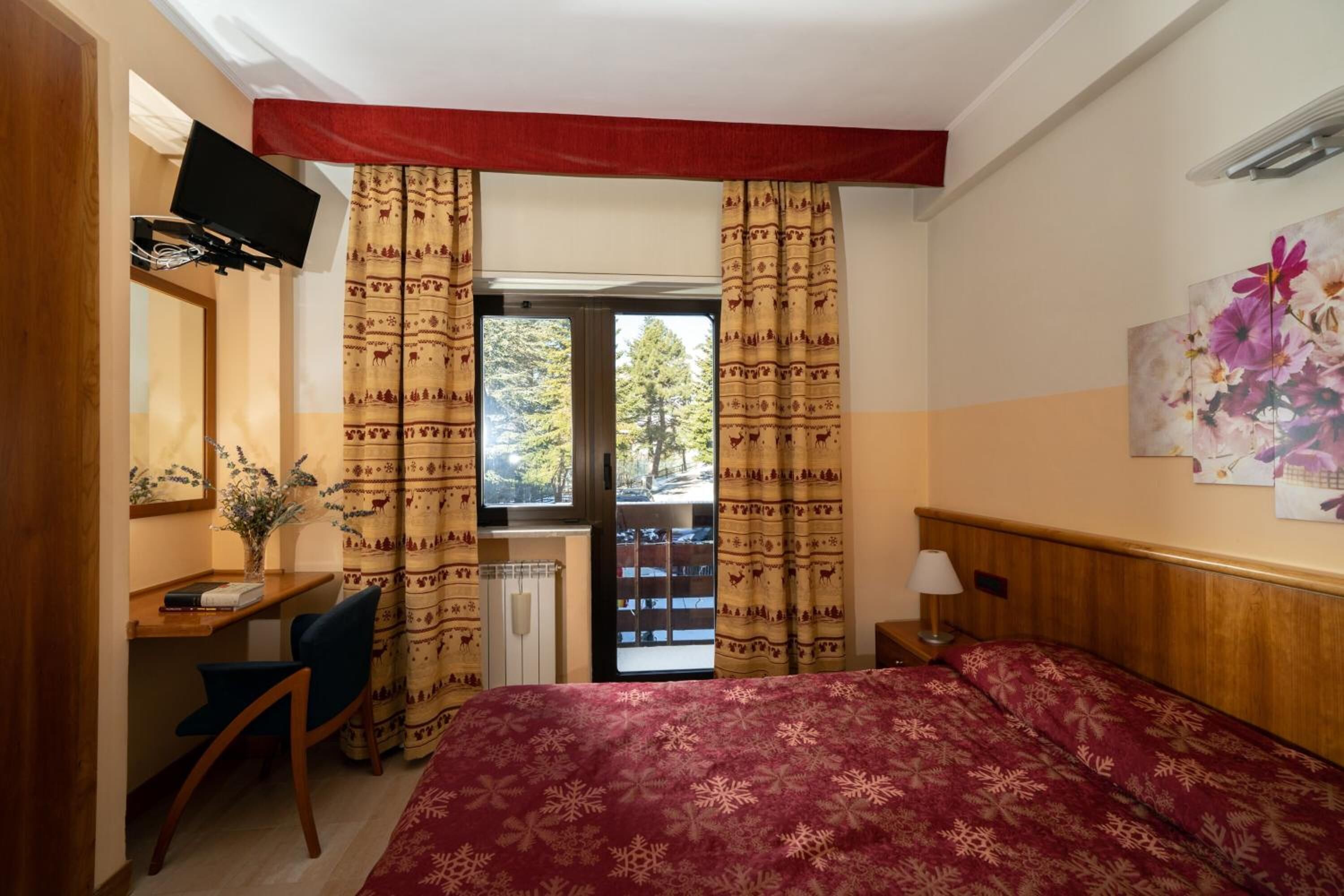 A hotel room with a bed, a desk, a chair, a TV mounted on the wall, and a window with curtains.