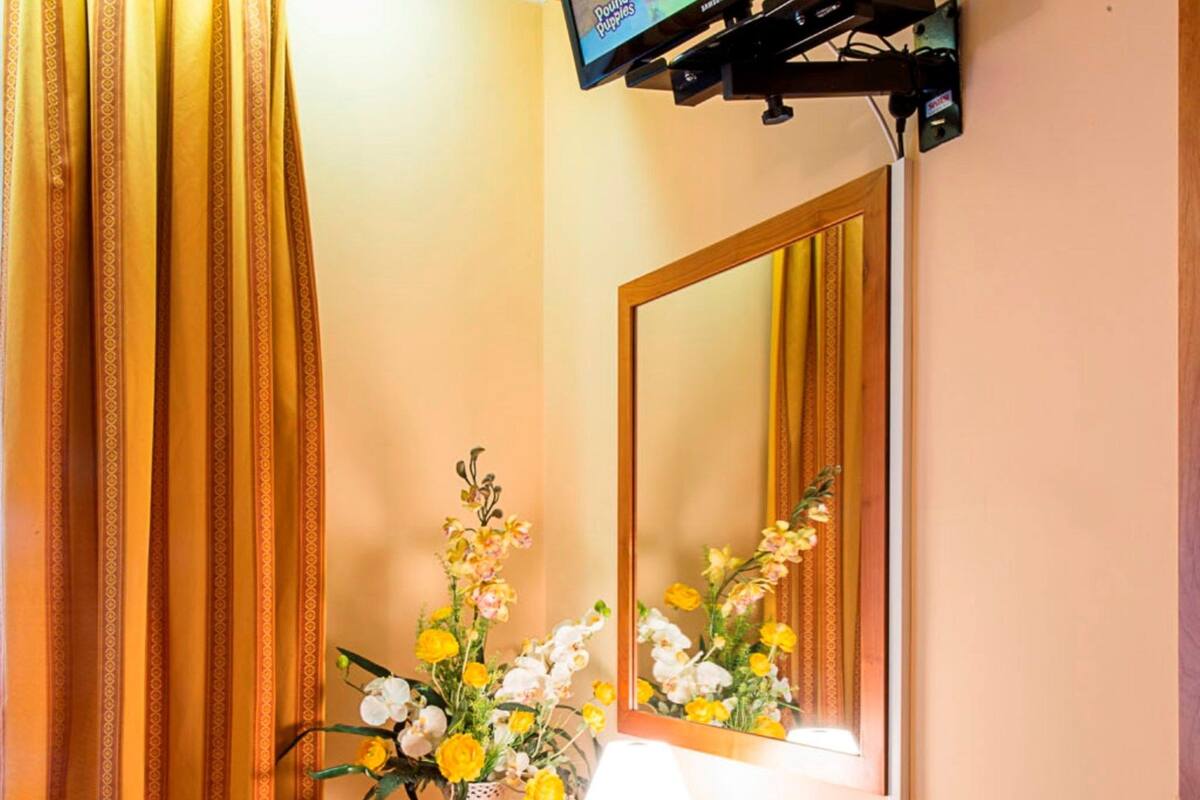 A hotel room with a TV mounted on the wall, a desk with a lamp, a vase of flowers, a chair, and a minibar.