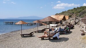 A beach with wooden loungers and thatched umbrellas, a rocky shoreline, and a view of the sea and distant mountains. - Assos Terrace Hotel (Ayvacik)