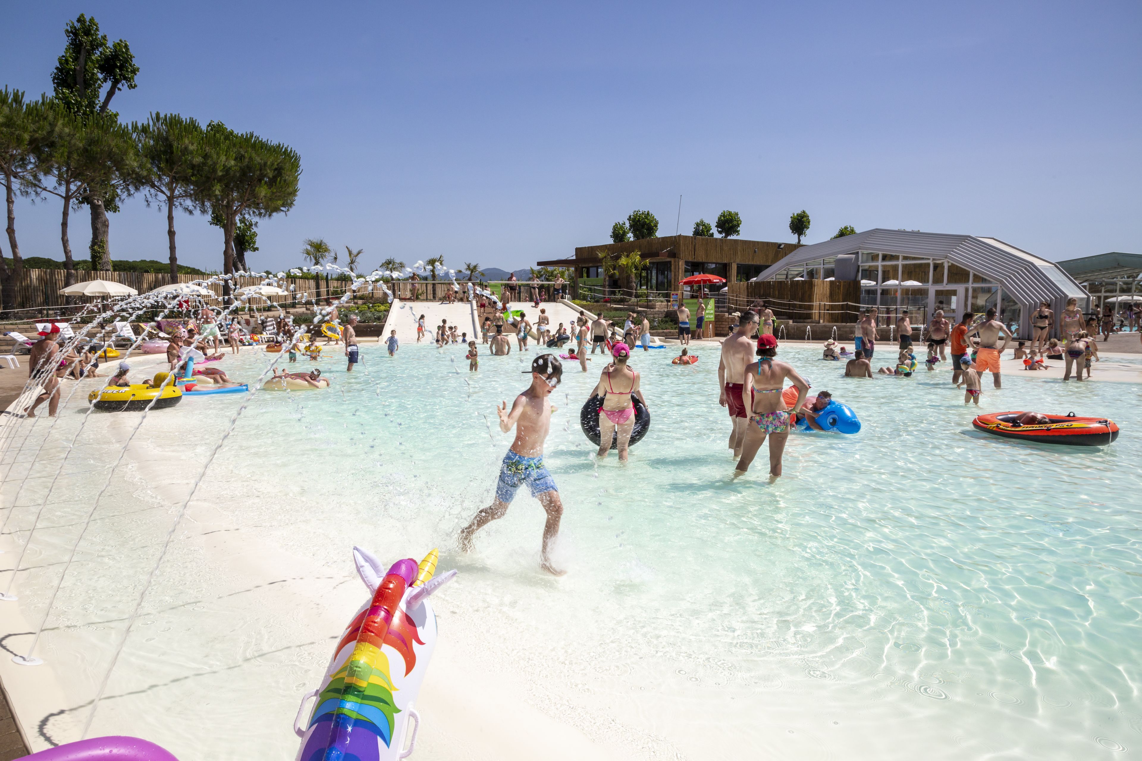 Un área de piscina concurrida, con gente nadando y jugando, rodeada de sombrillas y sillas de playa.