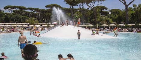 Una piscina con gente disfrutando del agua, rodeada de sombrillas y árboles.