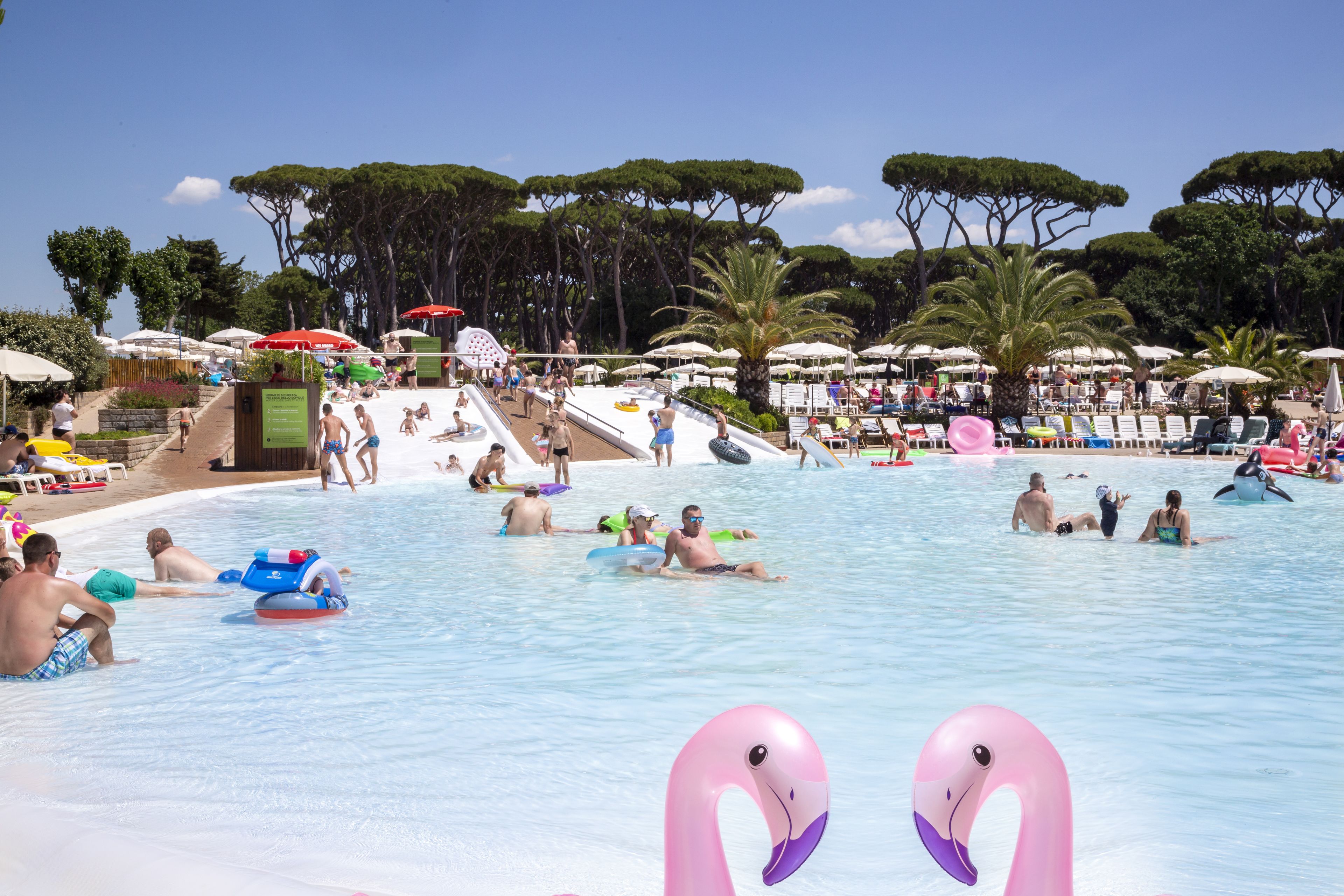 A swimming pool area with multiple people, inflatable toys, and umbrellas.